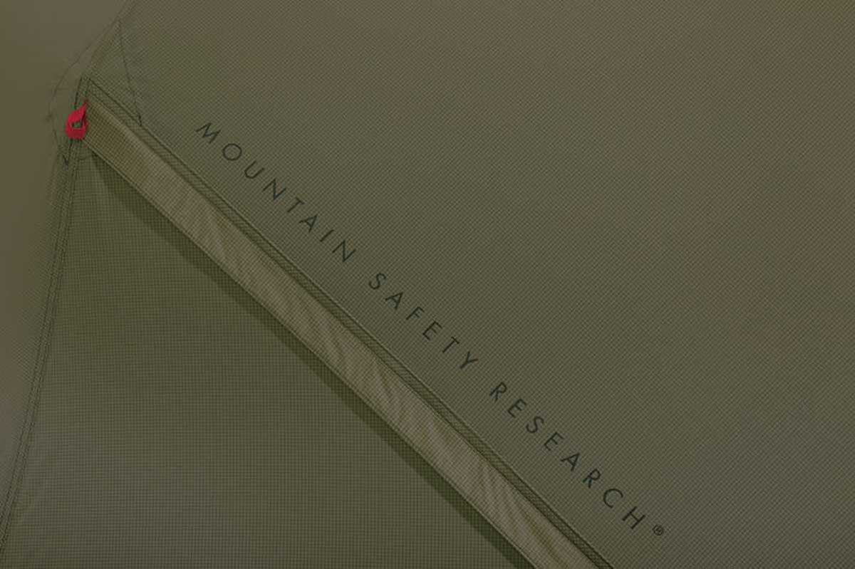 Närbild av grön textur på yttertältet med MOUNTAIN SAFETY RESEARCH-logga och röd detalj vid söm